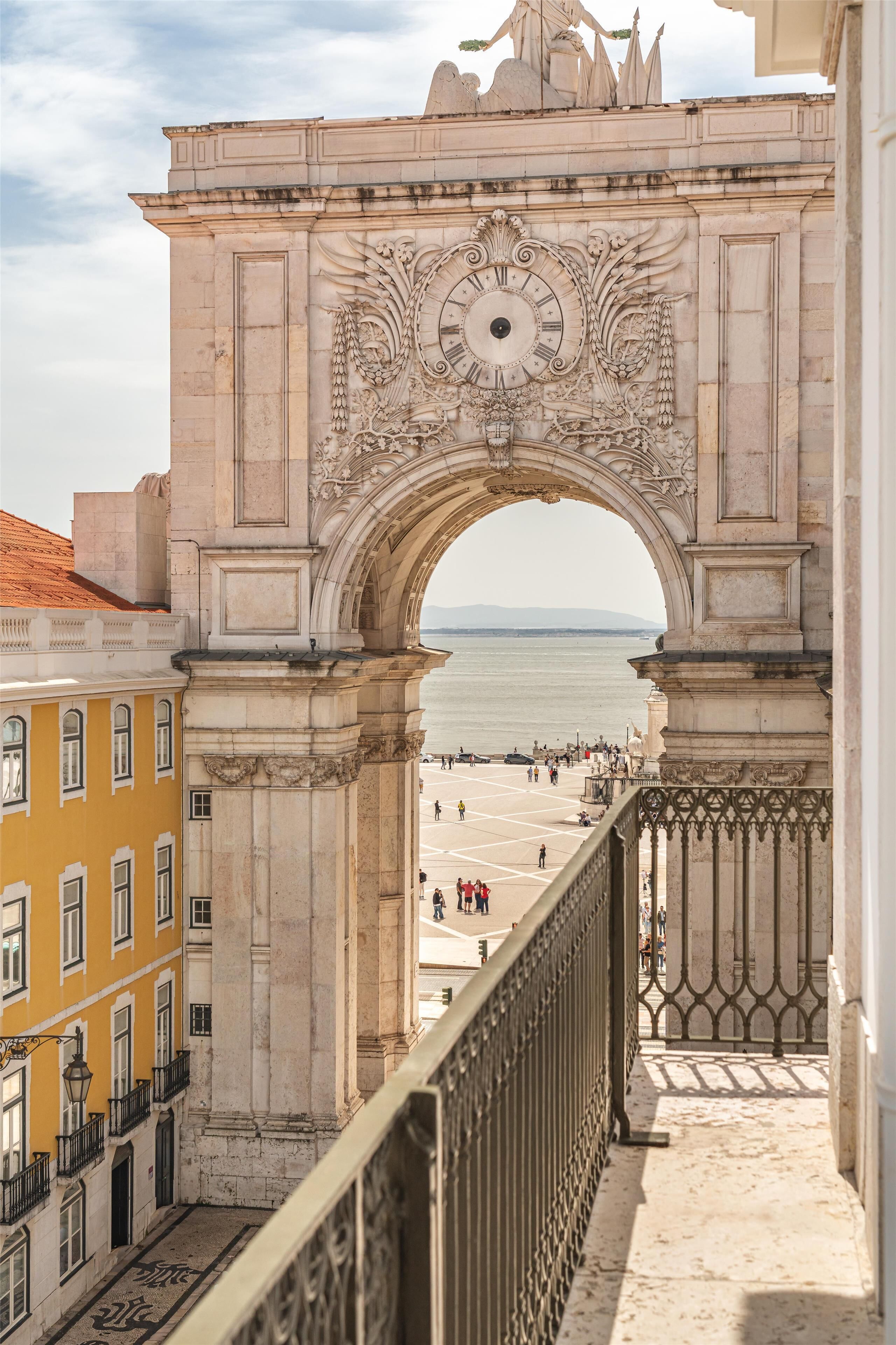 Un arco histórico con detalles tallados, con vista a una playa con gente y edificios.