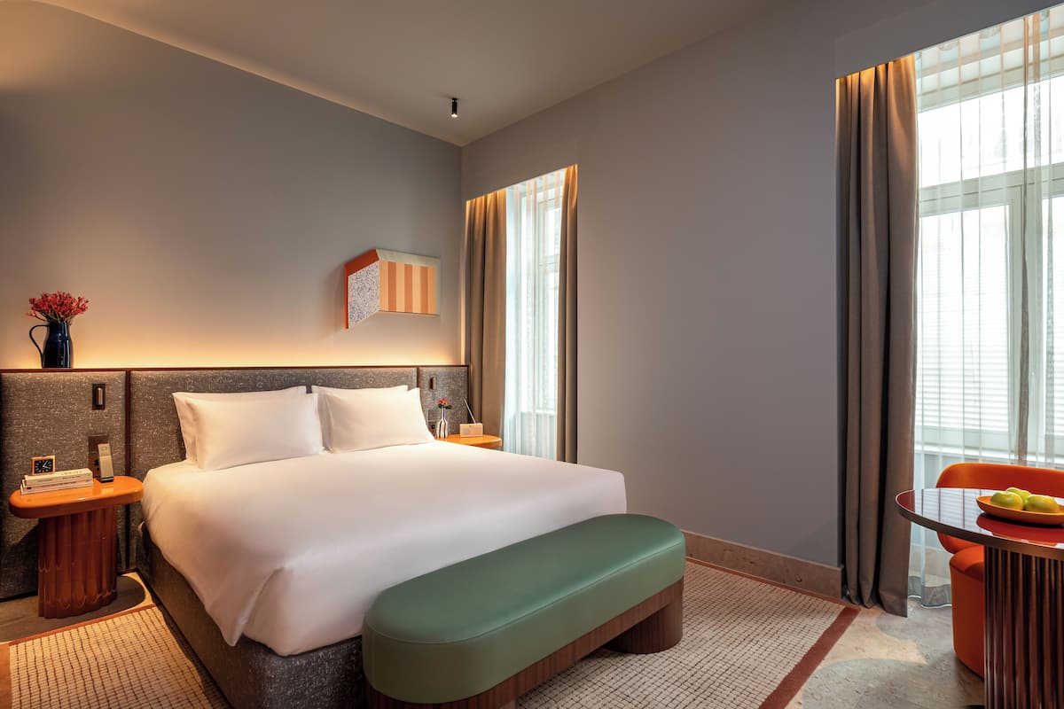 Habitación de hotel con una cama grande, una mesita de noche con lámpara, una silla y una pequeña mesa con fruta.