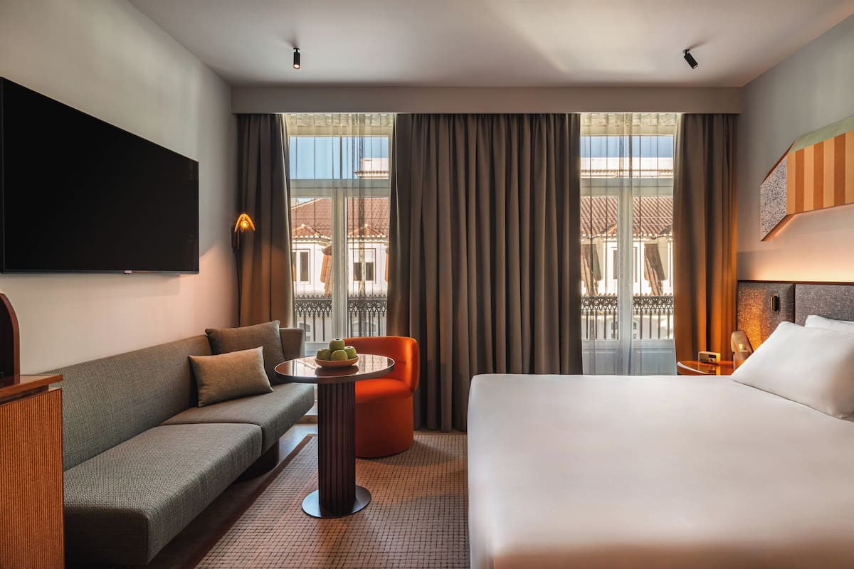 Habitación de hotel con sofá, cama, una pequeña mesa con fruta, televisión y balcón con cortinas.