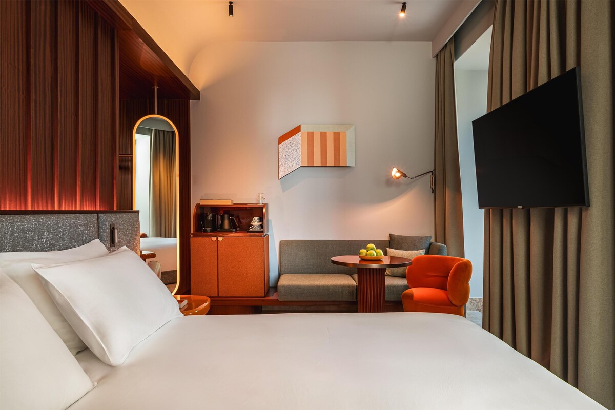 Habitación de hotel con cama, sofá, una mesita con fruta, televisor y espejo.