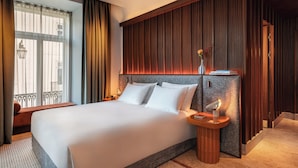 Una habitación de hotel moderna con una cama grande, cabecera de madera, mesita de noche con lámpara y vistas a la ciudad a través de la ventana.
