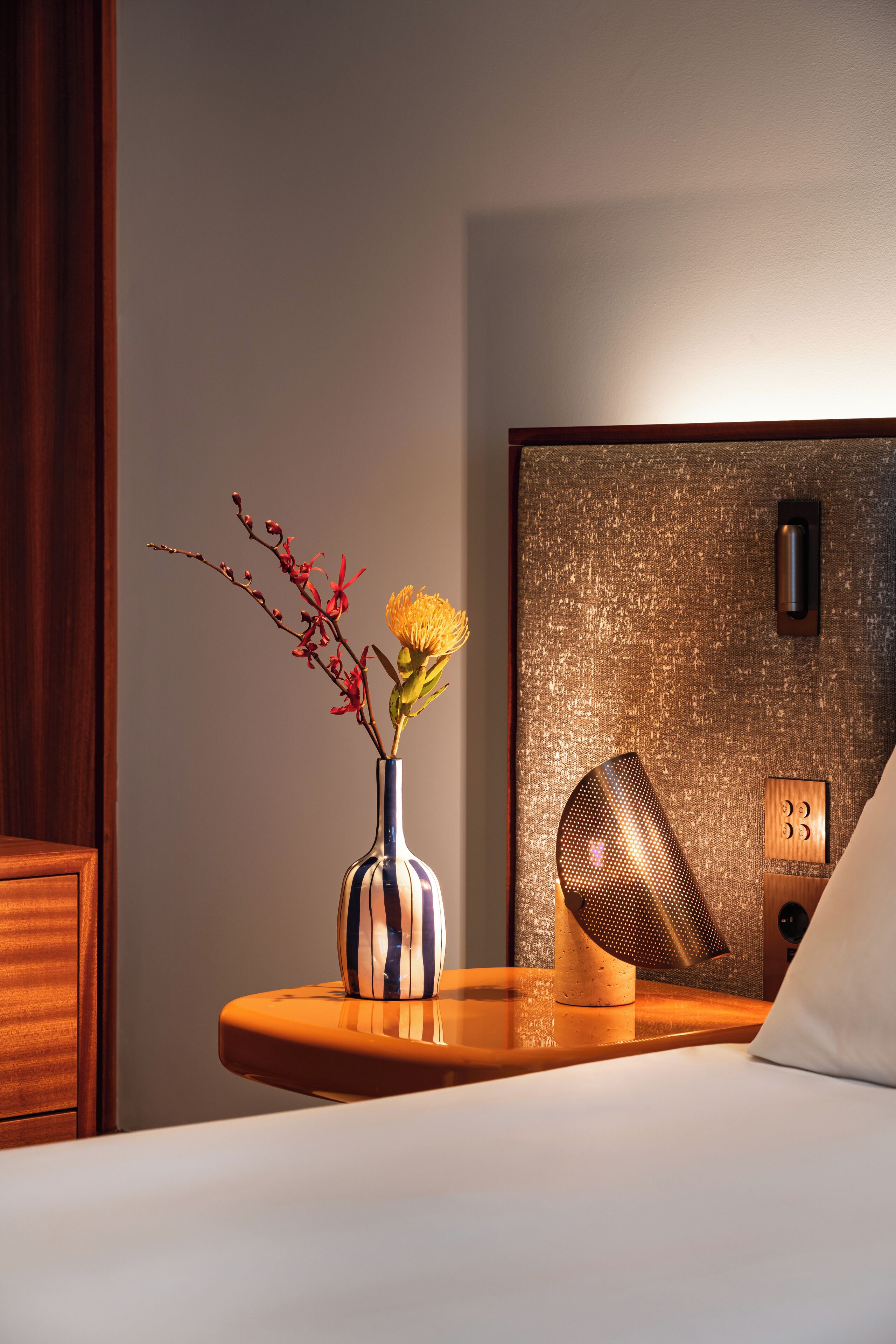 A hotel room with a bed, a bedside table with a vase of flowers, a wall-mounted lamp, and a textured wall panel.