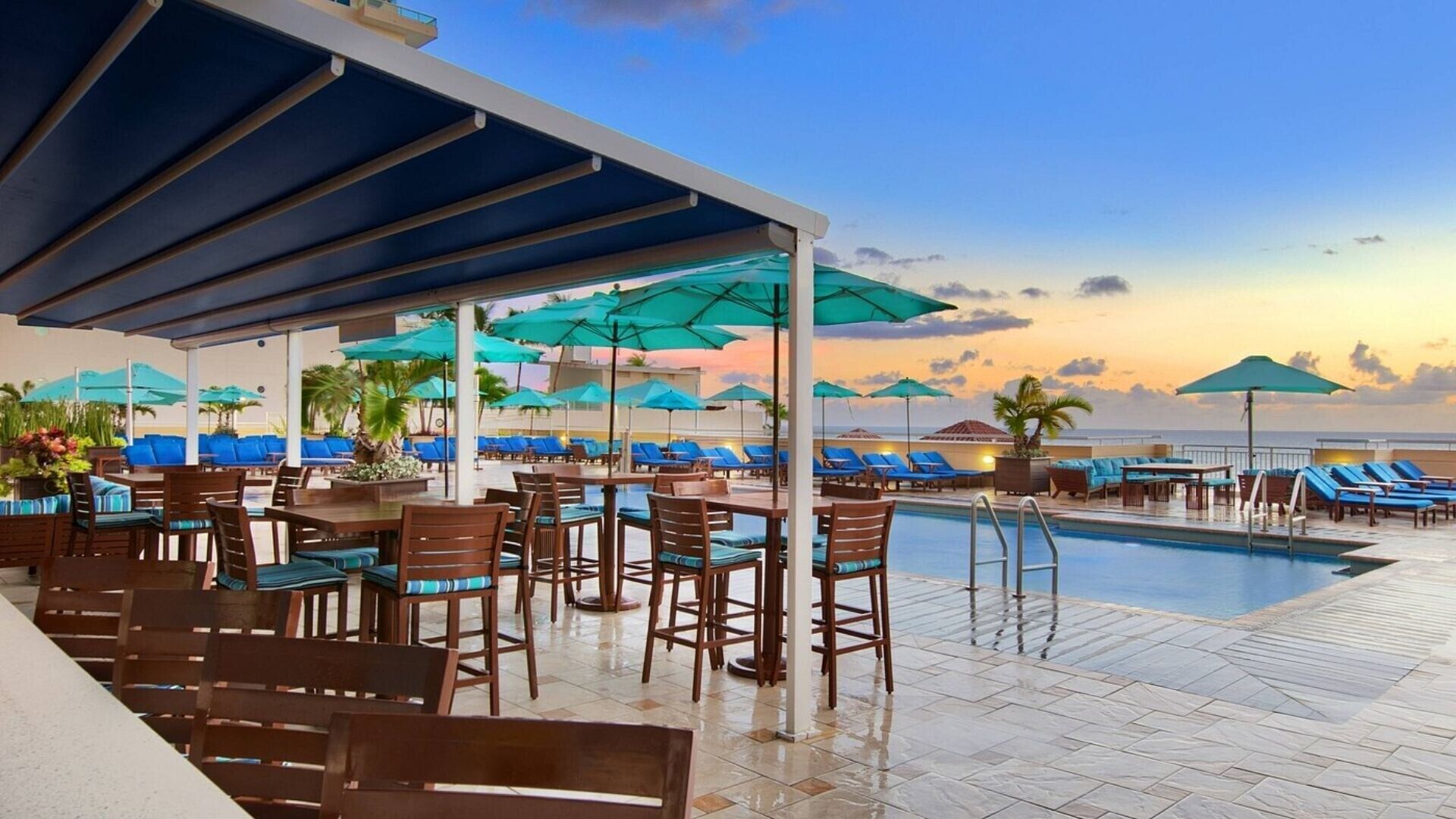 Área de piscina com mesas, cadeiras e guarda-sóis, com vista para o oceano ao pôr do sol.