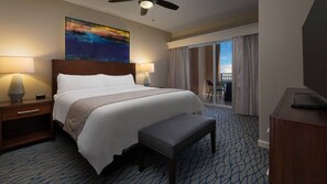 Una camera d'hotel con un letto grande, due lampade ai comodini, un ventilatore a soffitto, una televisione, un balcone con vista e un quadro appeso al muro.