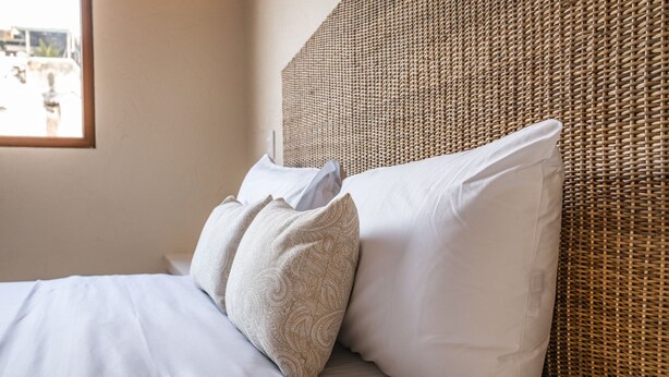 Una cama con sábanas blancas, almohadas estampadas, un cabecero tejido y una ventana con vistas al exterior.