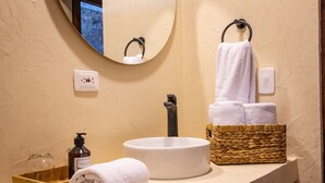 Une salle de bain avec un miroir rond, un lavabo blanc, une serviette, un flacon de lotion et un panier en osier.
