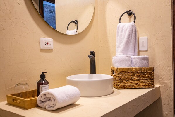 A bathroom with a round mirror, a white sink, a towel, a bottle of lotion, and a wicker basket.