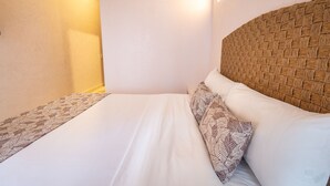 A neatly made bed with white linens and patterned pillows, a woven headboard, and a wall-mounted light fixture.