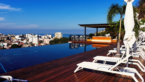 A rooftop infinity pool with wooden decking and white lounge chairs, overlooking a cityscape and the sea. - Oasis of Beauty (Puerto Vallarta)