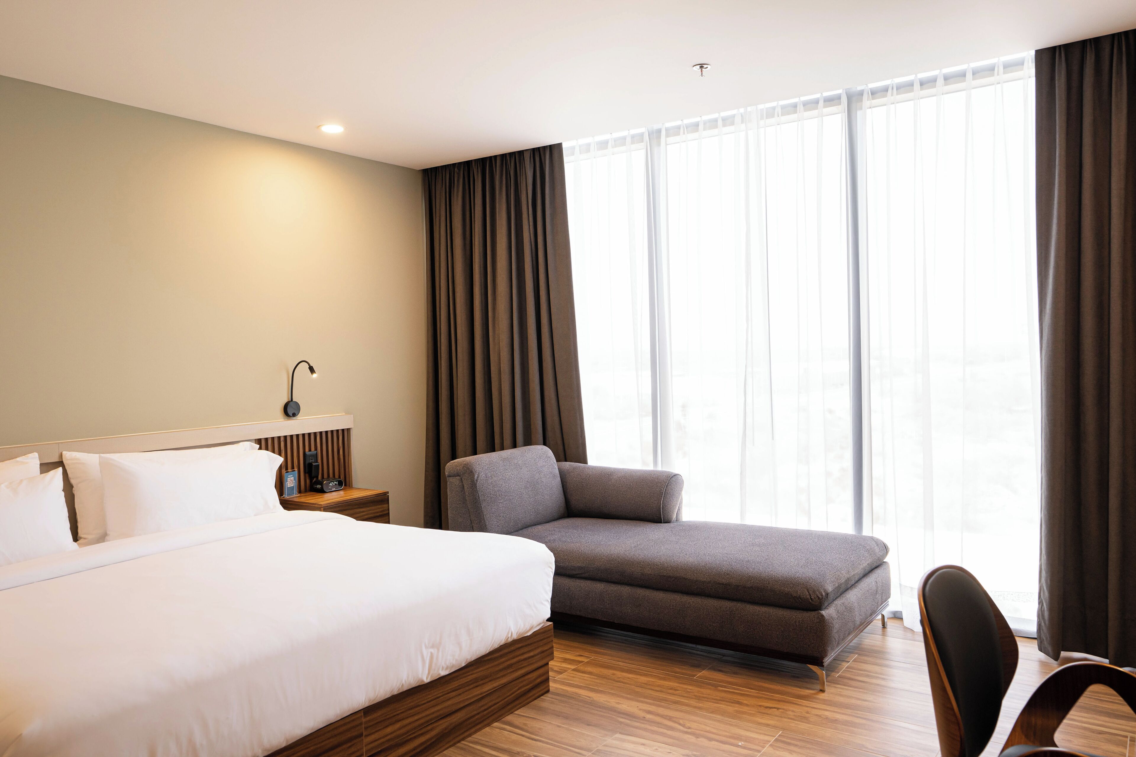 Una habitación de hotel moderna con una cama grande, un sofá, un escritorio con lámpara y un ventanal con cortinas.