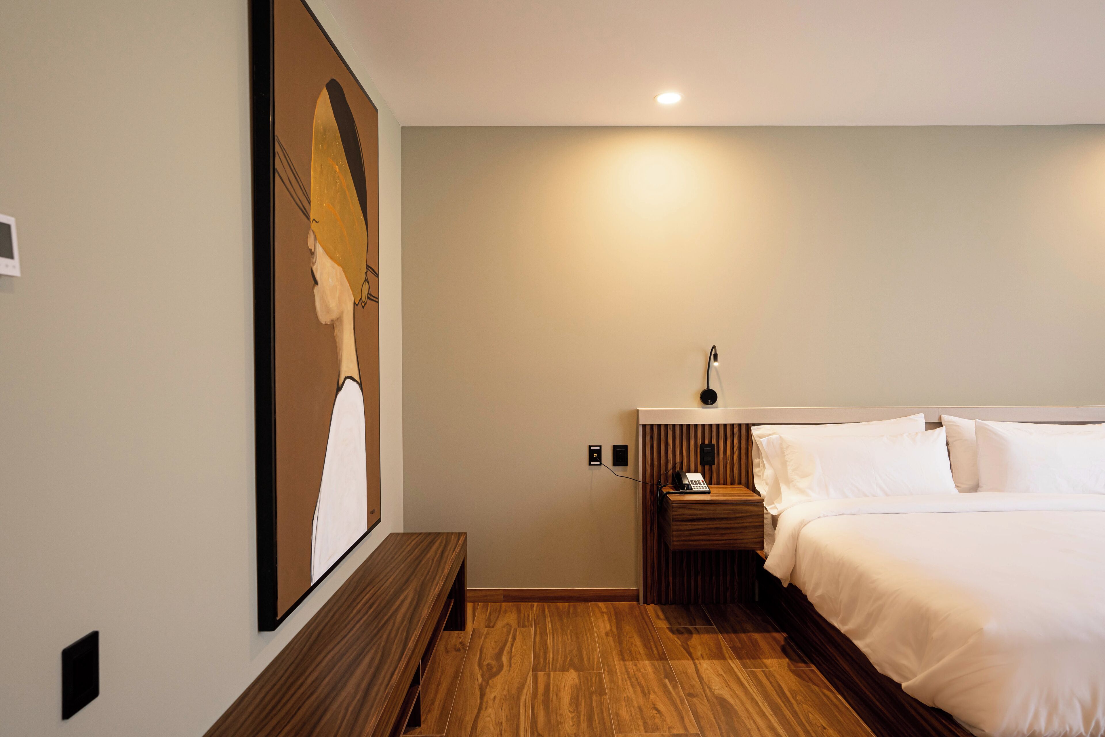 Ein Hotelzimmer mit Bett, Nachttisch mit Lampe, einer Holzbank und einem Wandbild.