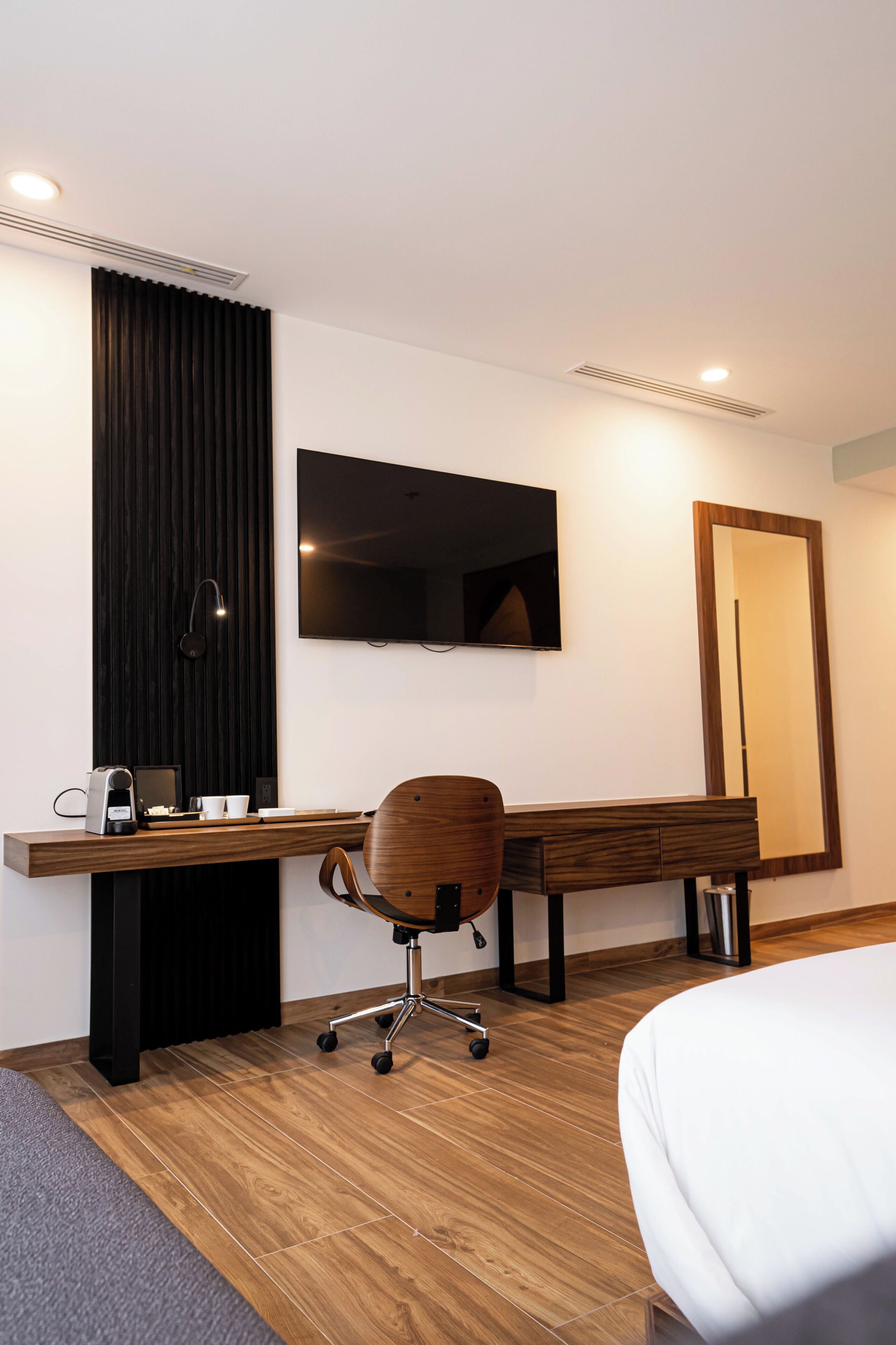 Habitación de hotel moderna con un escritorio de madera, una silla, una cama, un televisor de pantalla plana y un espejo grande.