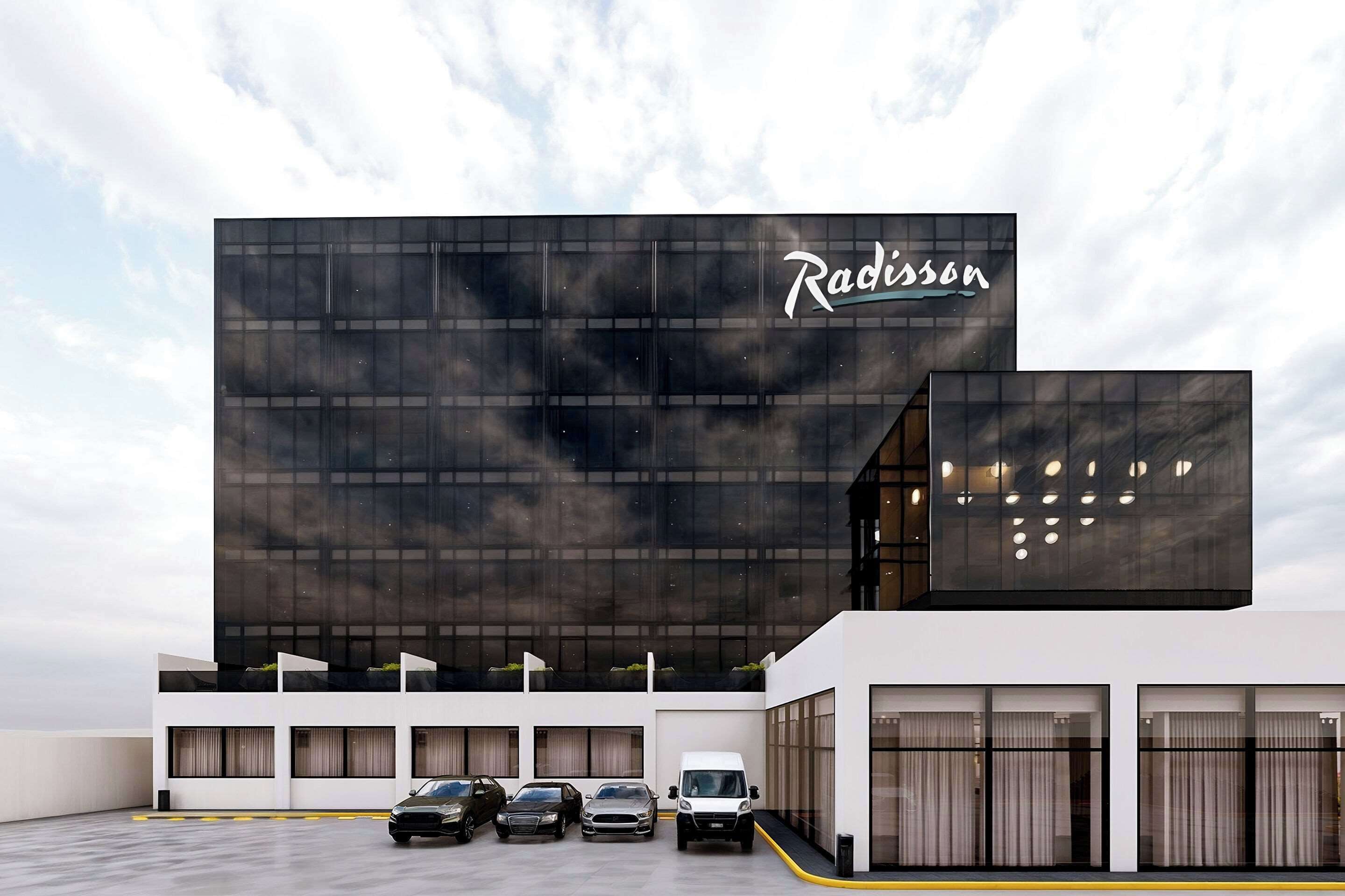 A modern building with the name 'Radisson' on the facade. There are multiple cars parked in front, and the building has large windows and a flat roof.