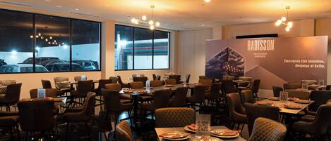 A restaurant with multiple tables and chairs, large windows, and a modern chandelier.