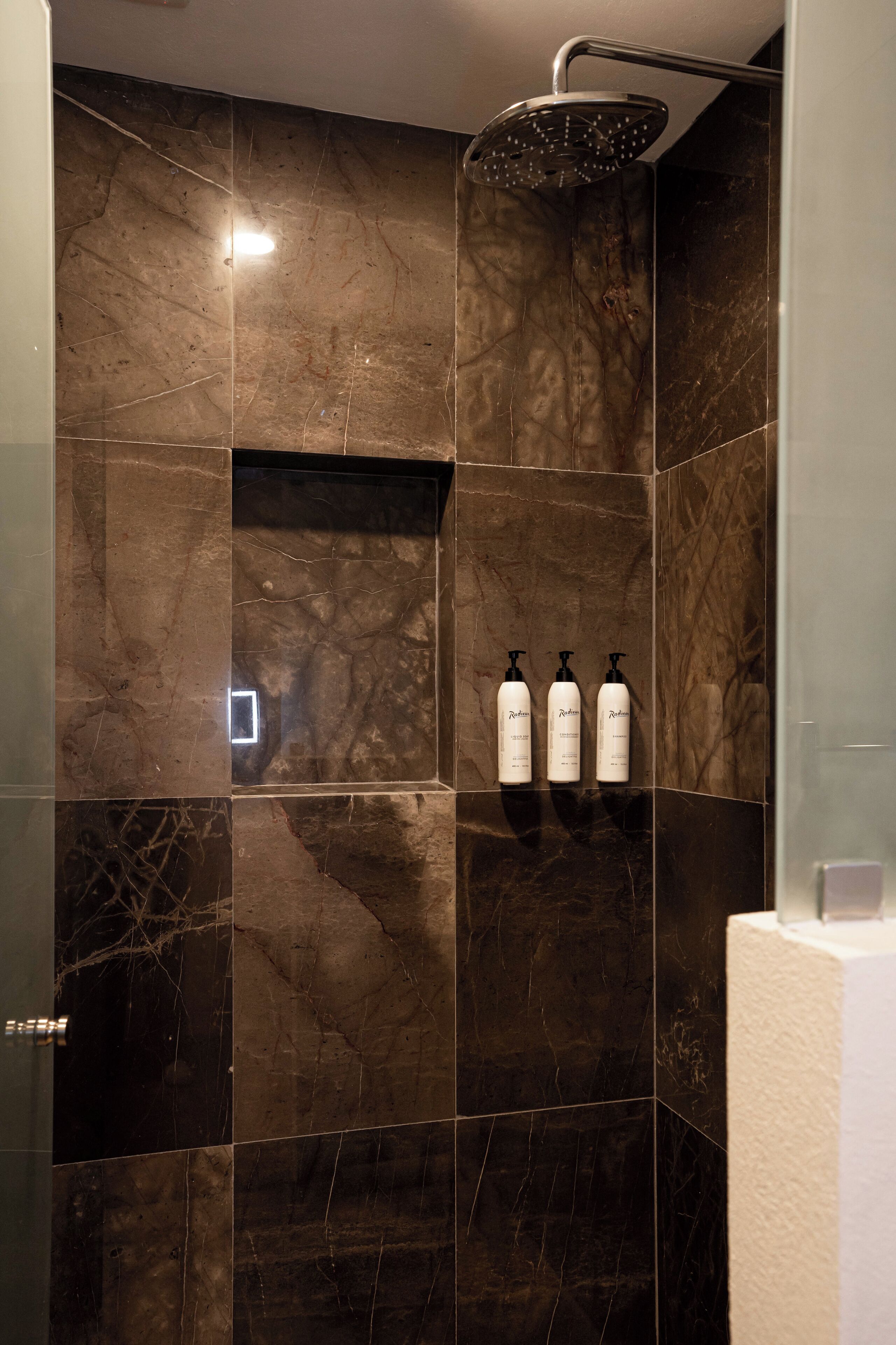 A modern bathroom with dark marble tiles, a showerhead, and three bottles of shampoo.