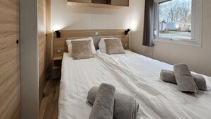 Een net opgemaakt bed met witte lakens en twee kussens, een houten hoofdbord en een raam met uitzicht op een huis en bomen.