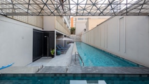 A swimming pool with a geometric ceiling structure, lounge chairs, and a door leading to a covered area.