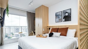 A modern hotel room with a large bed, a small table with breakfast, a city view, and two framed pictures on the wall.