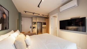A modern hotel room with a large bed, a flat-screen TV mounted on the wall, a kitchenette, and a door leading to a bathroom.