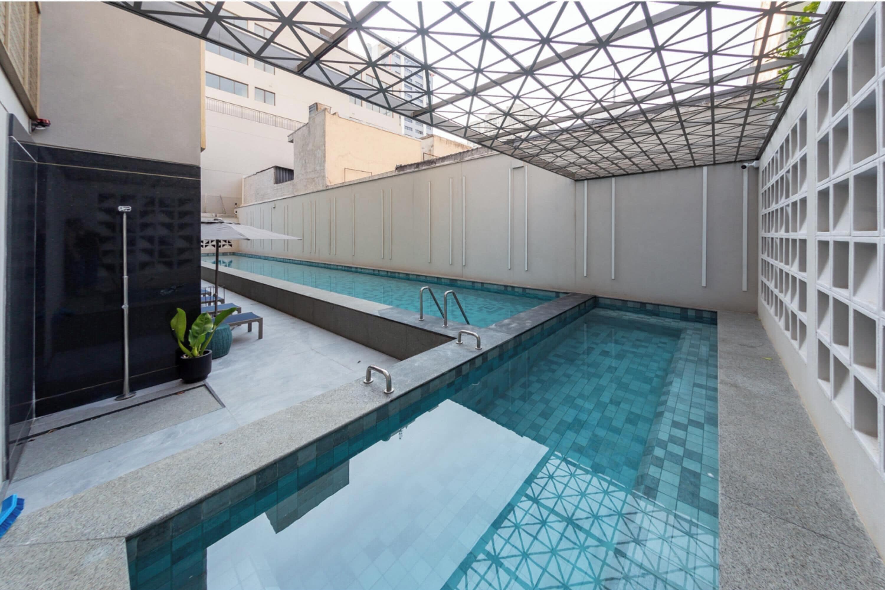 Una piscina interior moderna con techo de vidrio geométrico, suelo de baldosas y un pequeño rincón de descanso con una planta en maceta.