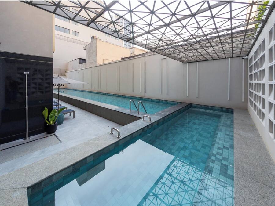 Una piscina interior moderna con techo de vidrio geométrico, suelo de baldosas y un pequeño rincón de descanso con una planta en maceta.
