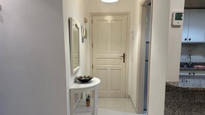 Un couloir avec une porte blanche, une petite table sur laquelle se trouvent un bol et une plante, et un luminaire au plafond.