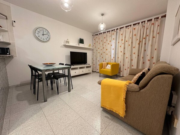 A modern living room with a sofa, a dining table with chairs, a TV, a clock, and a curtain with a floral pattern. - Casa Cabana - El Médano (Granadilla de Abona)