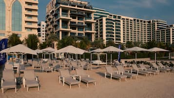 A beach with sun loungers and umbrellas, a modern hotel building, and palm trees.