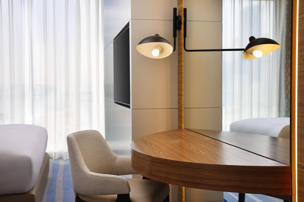 A hotel room with a round wooden table, two chairs, a flat-screen TV mounted on the wall, and a modern pendant light fixture.