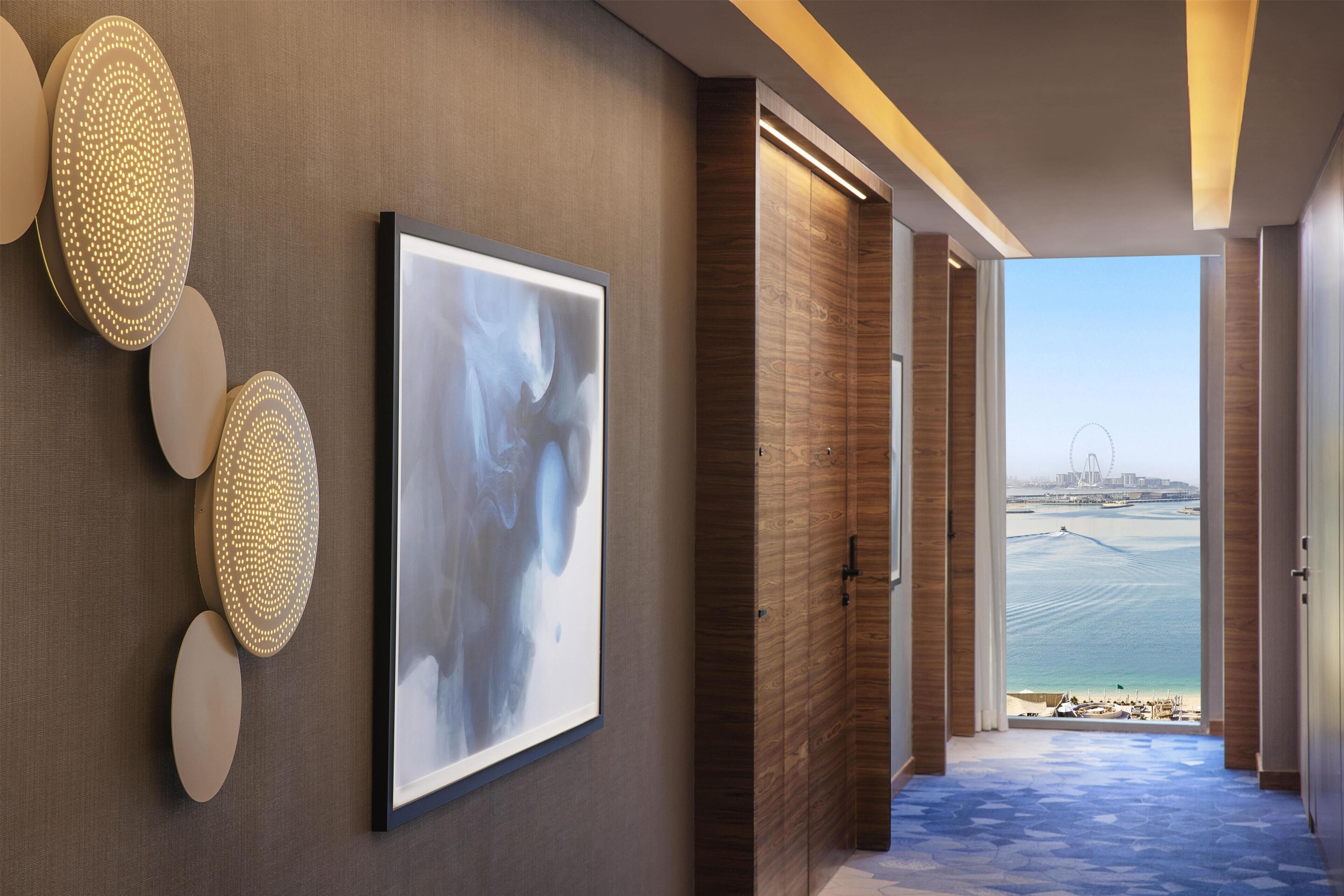 A hallway with a framed abstract painting, illuminated wall decor, and a view of the sea and a bridge.