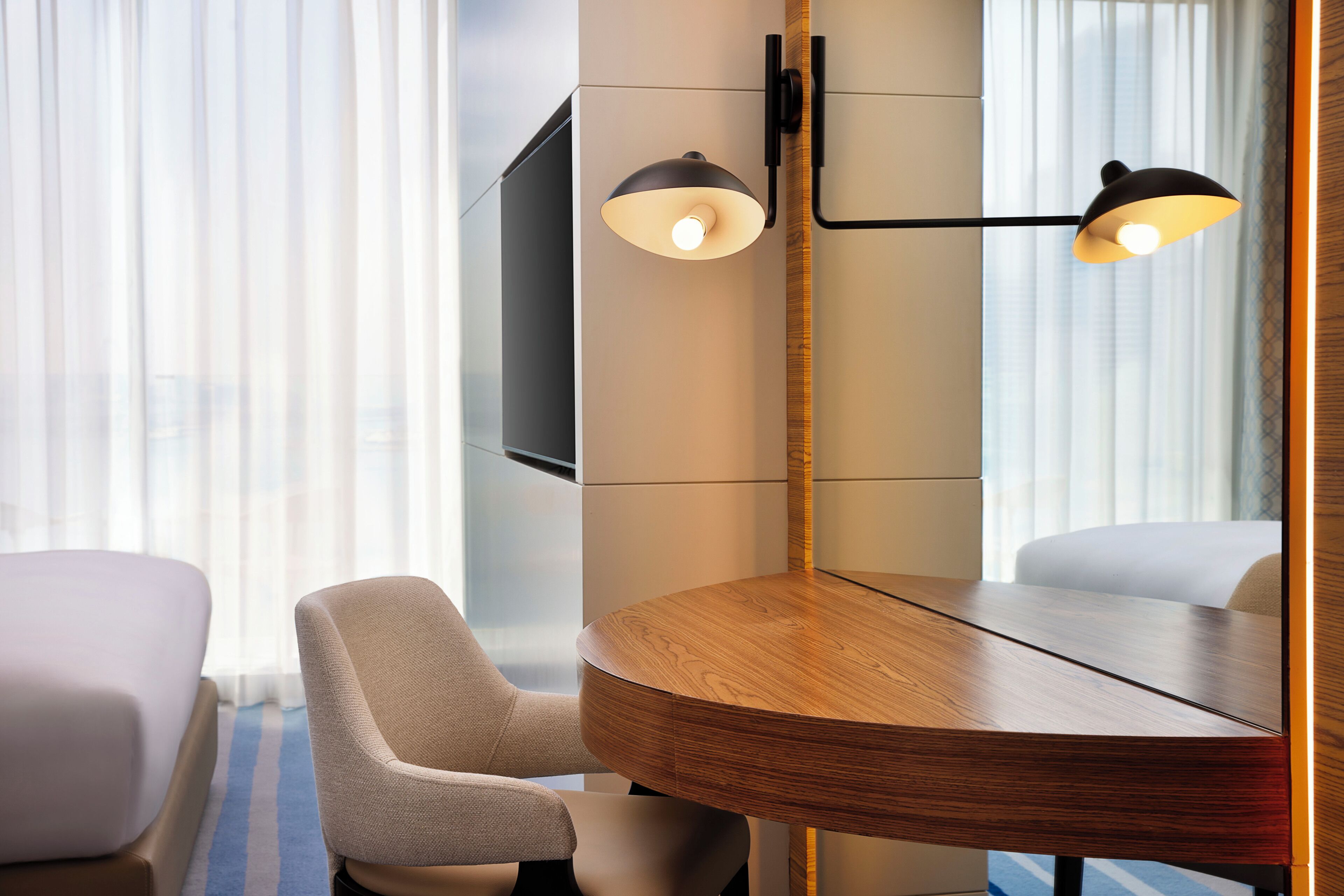 Habitación de hotel con una mesa redonda de madera, dos sillas, un televisor de pantalla plana montado en la pared y un ventanal con cortinas de gasa.