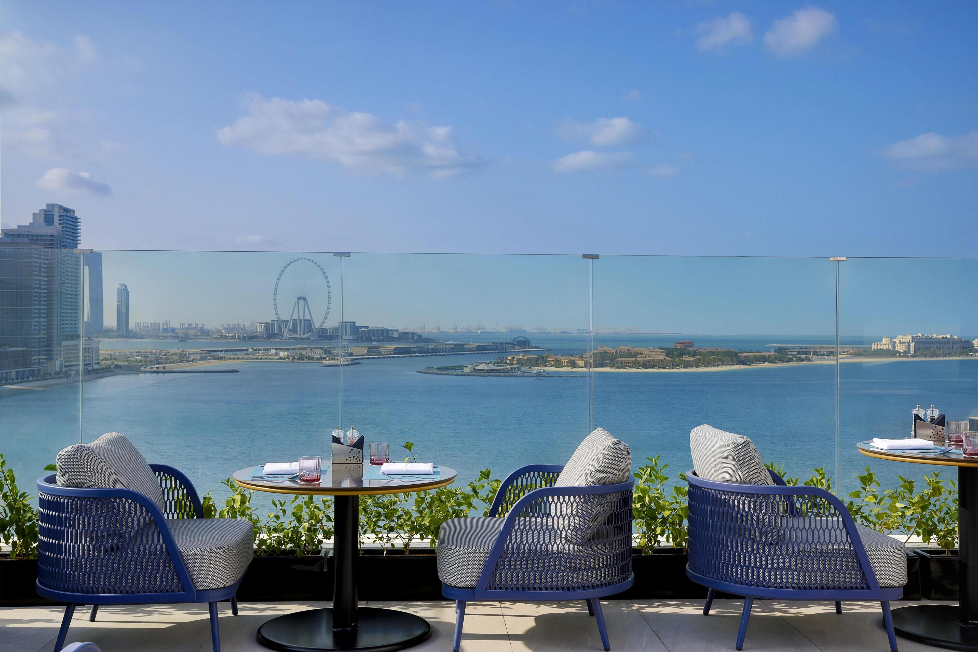 A balcony with two chairs and a table, overlooking a body of water with a distant cityscape and a Ferris wheel.