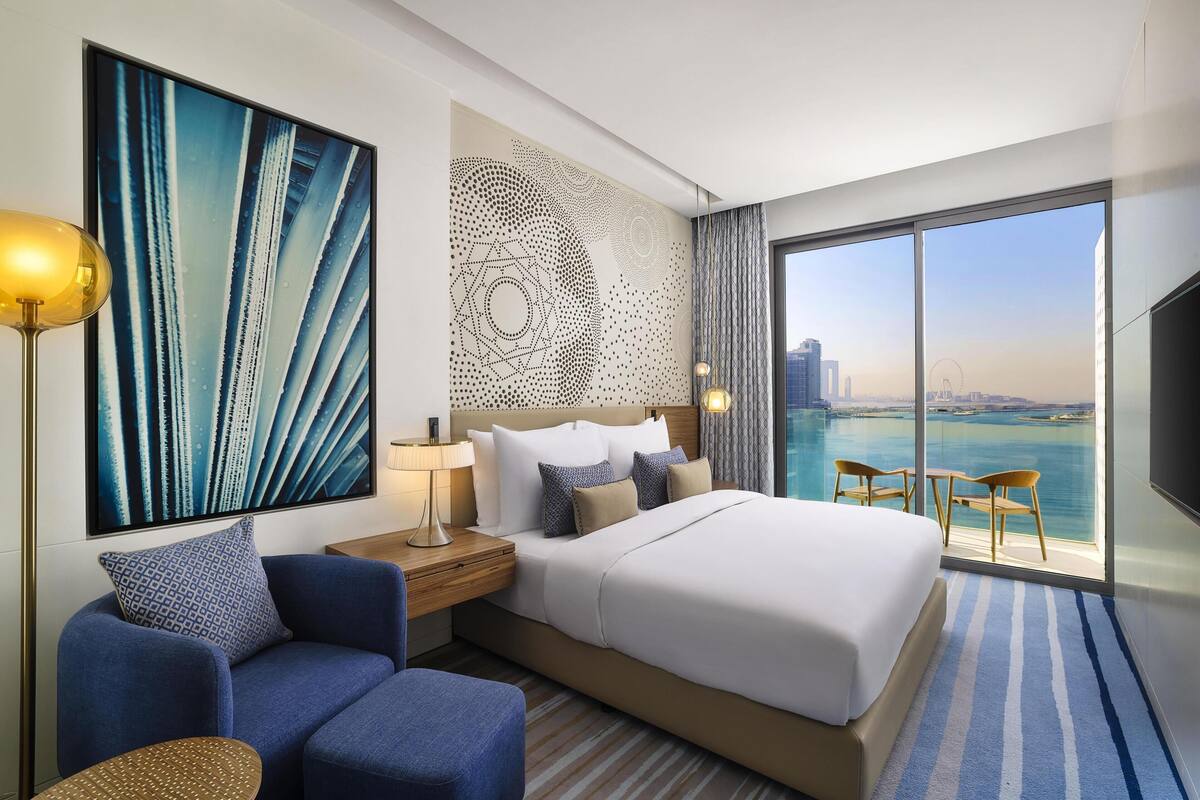 A modern hotel room with a large bed, a seating area, a view of the water, and a decorative wall.