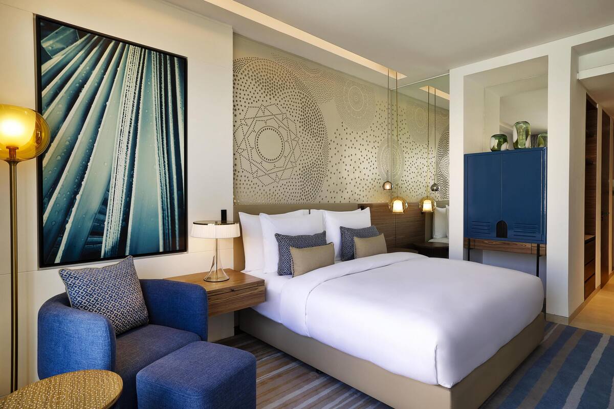 A modern hotel room with a large bed, a seating area with a blue sofa, a side table with a lamp, and a wall-mounted artwork.