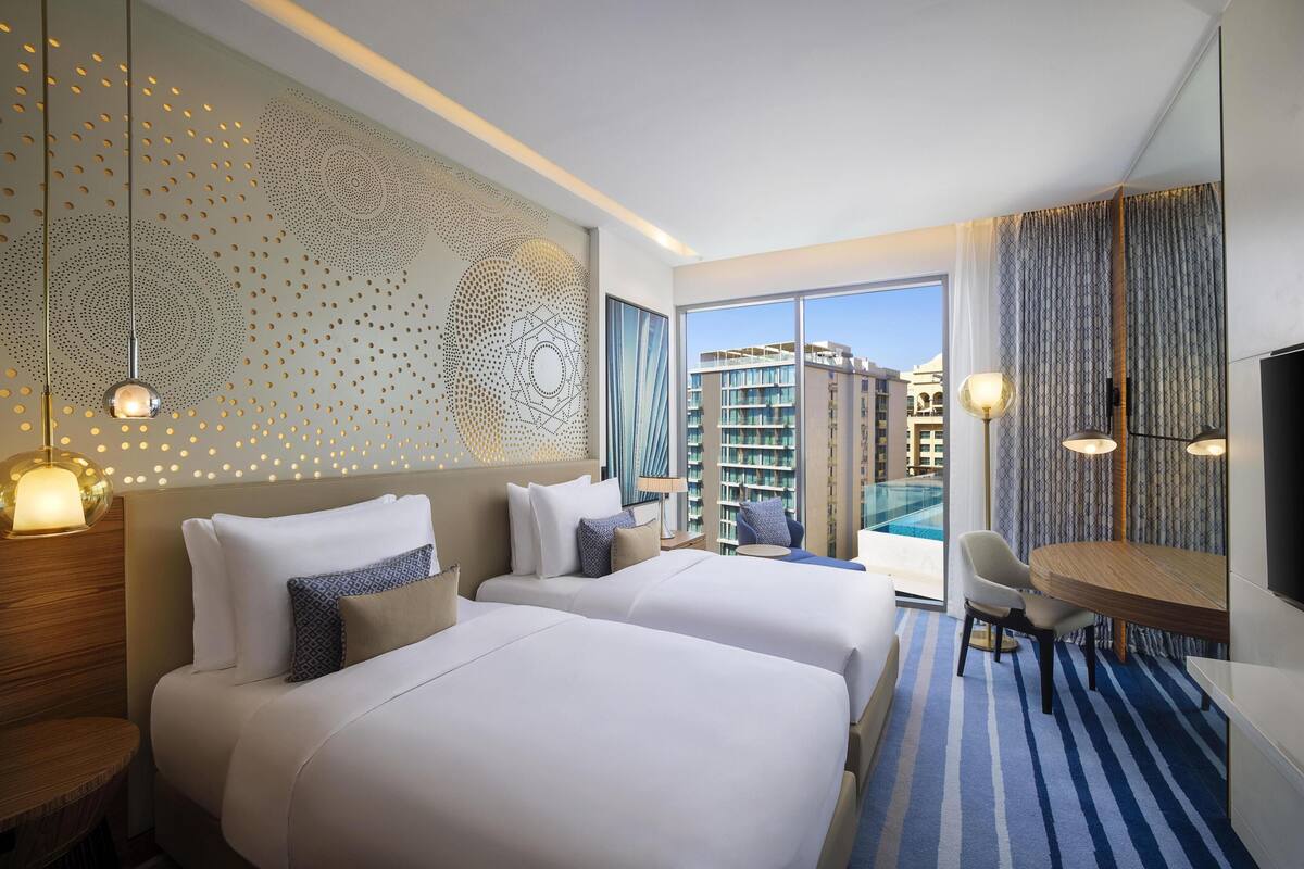 A hotel room with two beds, a dining table, a TV, and a view of a building.