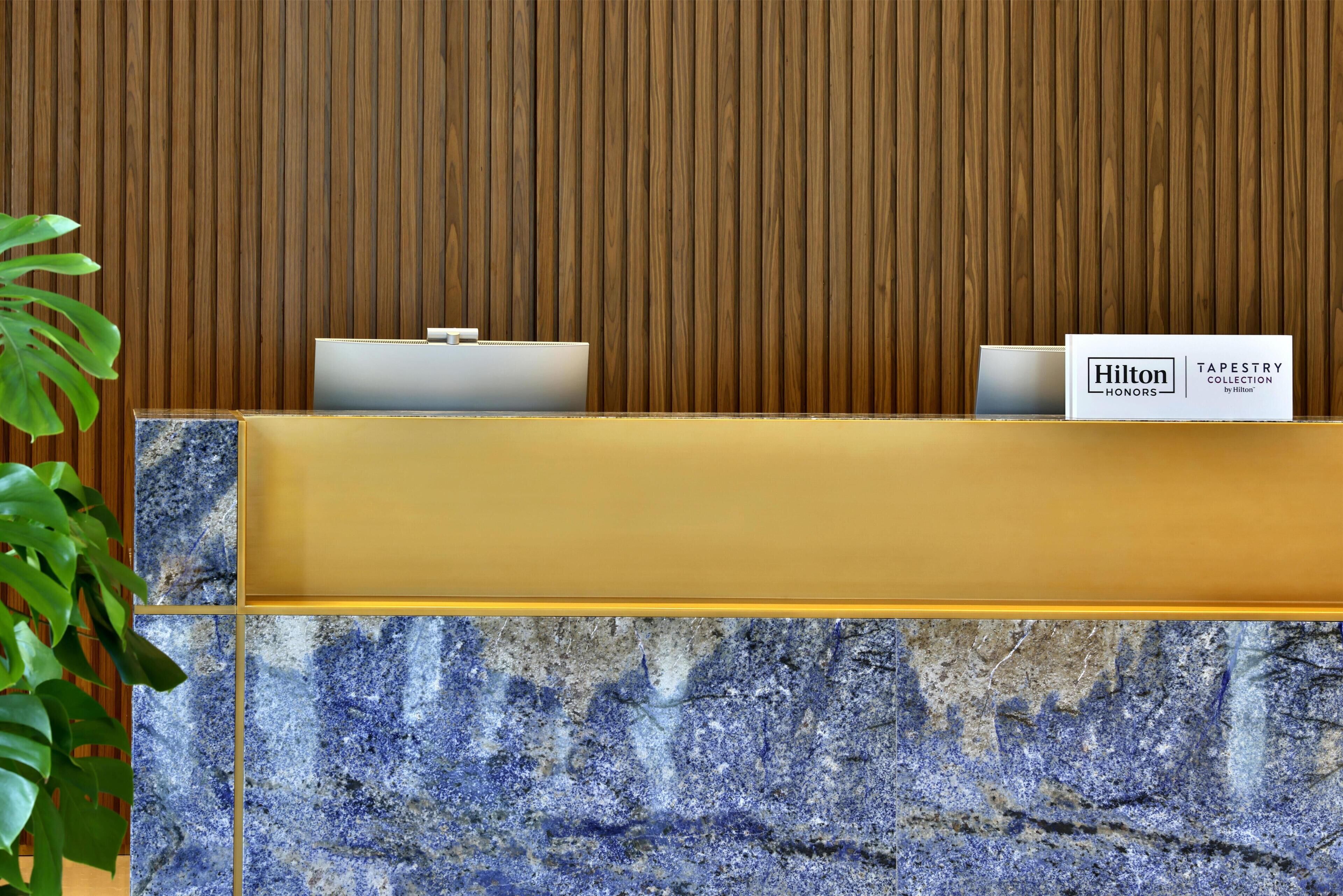 Un comptoir d’accueil avec un panneau en bois en arrière-plan, une enseigne indiquant « Hilton Honors Tapestry Collection by Hilton », et une plante en pot placée à gauche.