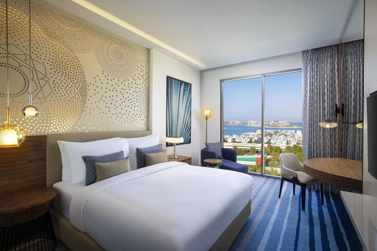 A hotel room with a large bed, a desk, a chair, a view of the city, and a large window.