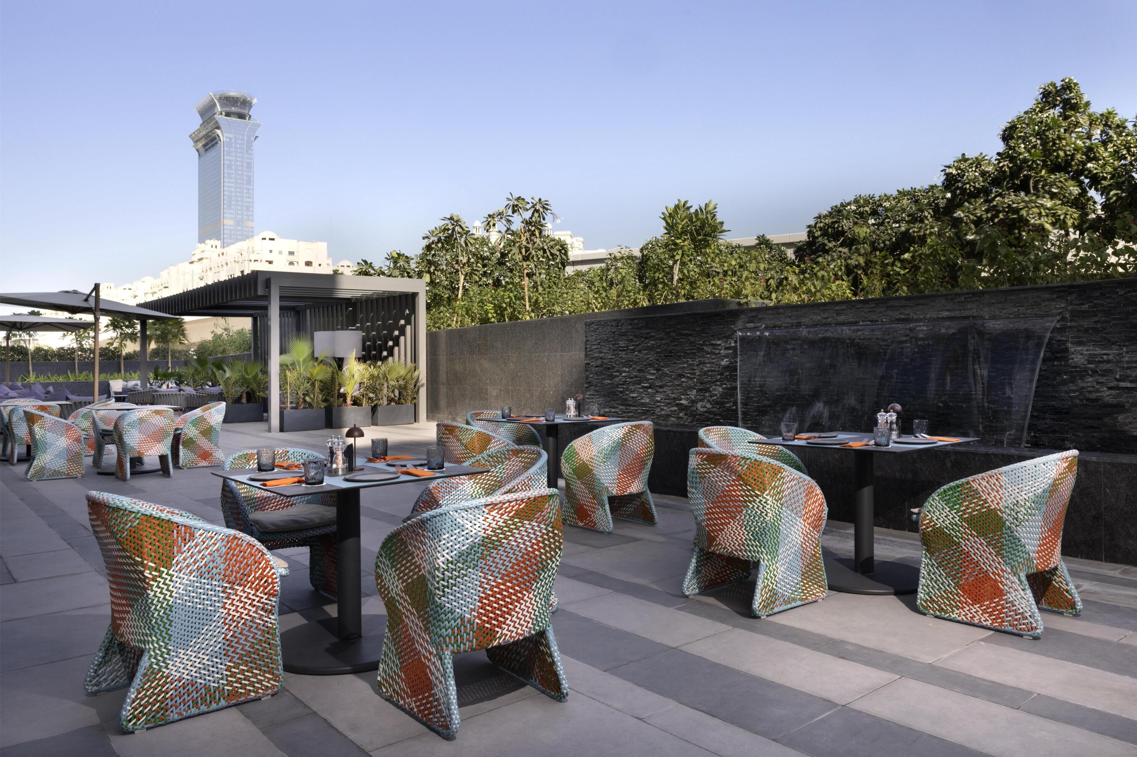 Espace extérieur aménagé avec des chaises et des tables aux motifs originaux, un mur en pierre et une vue sur un grand immeuble en arrière-plan.