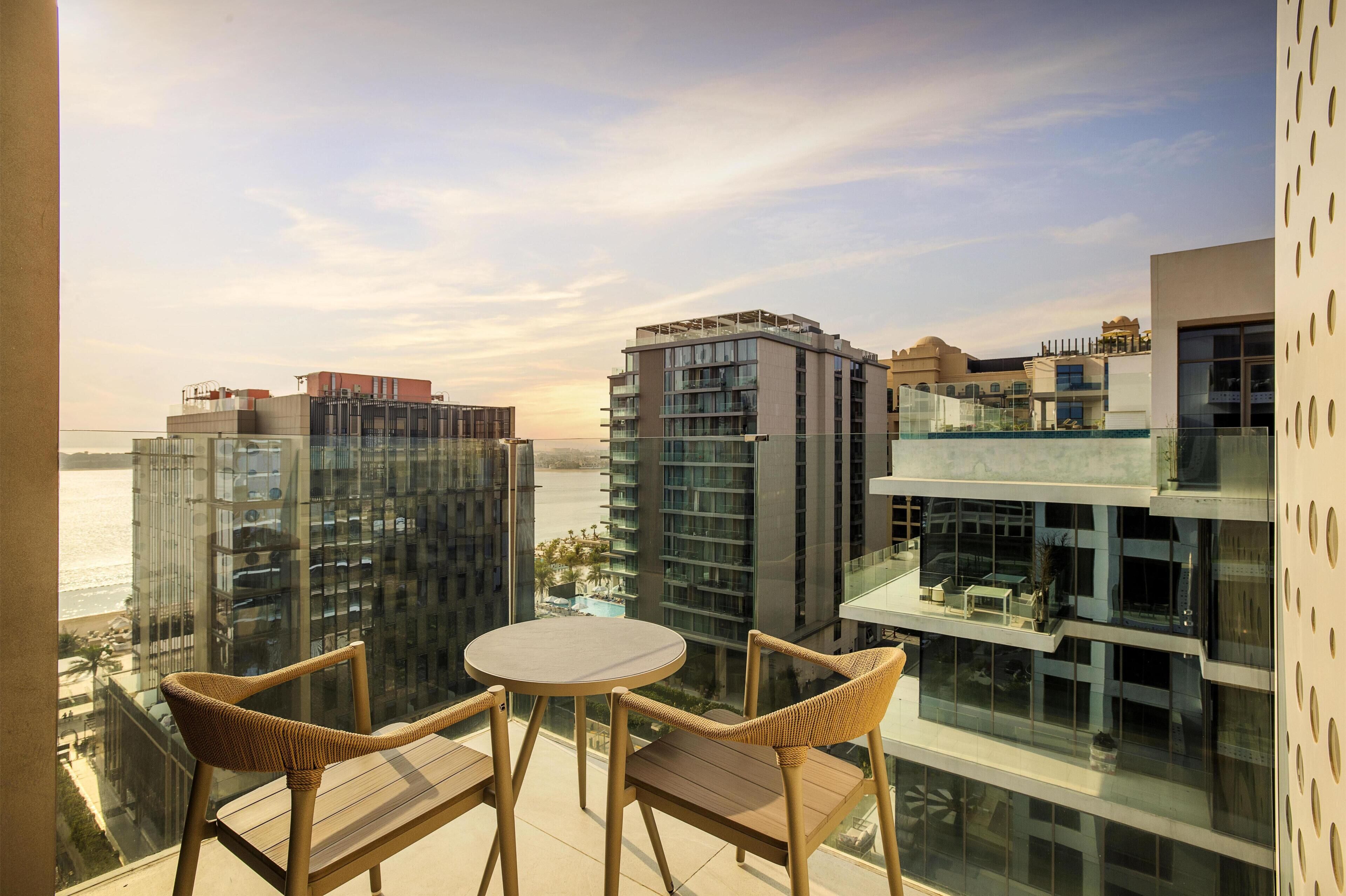 Un balcón con dos sillas de mimbre y una mesita pequeña, con vistas a un moderno complejo de edificios y a la playa.