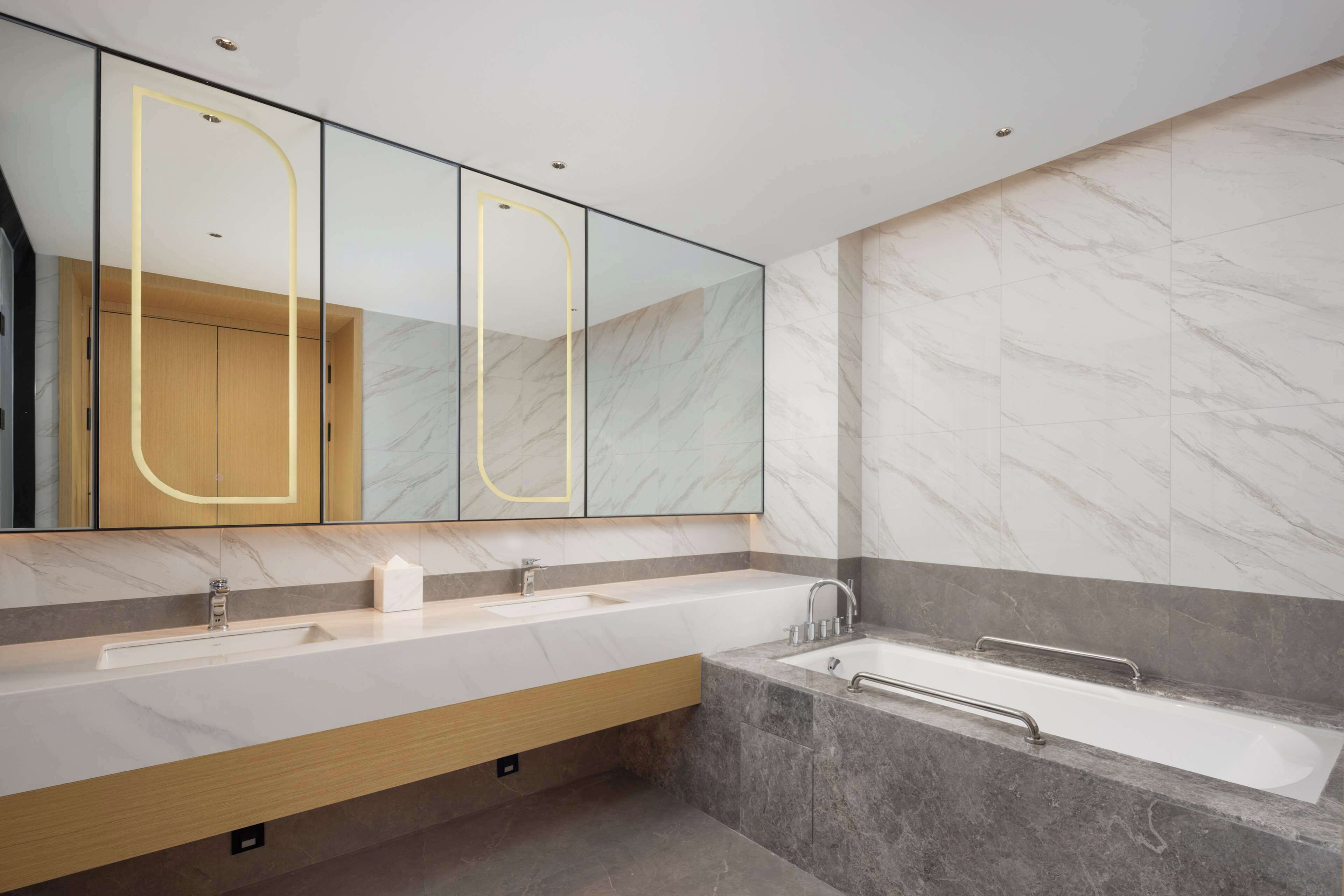 A modern bathroom with a large mirror, two sinks, a bathtub, and marble walls.