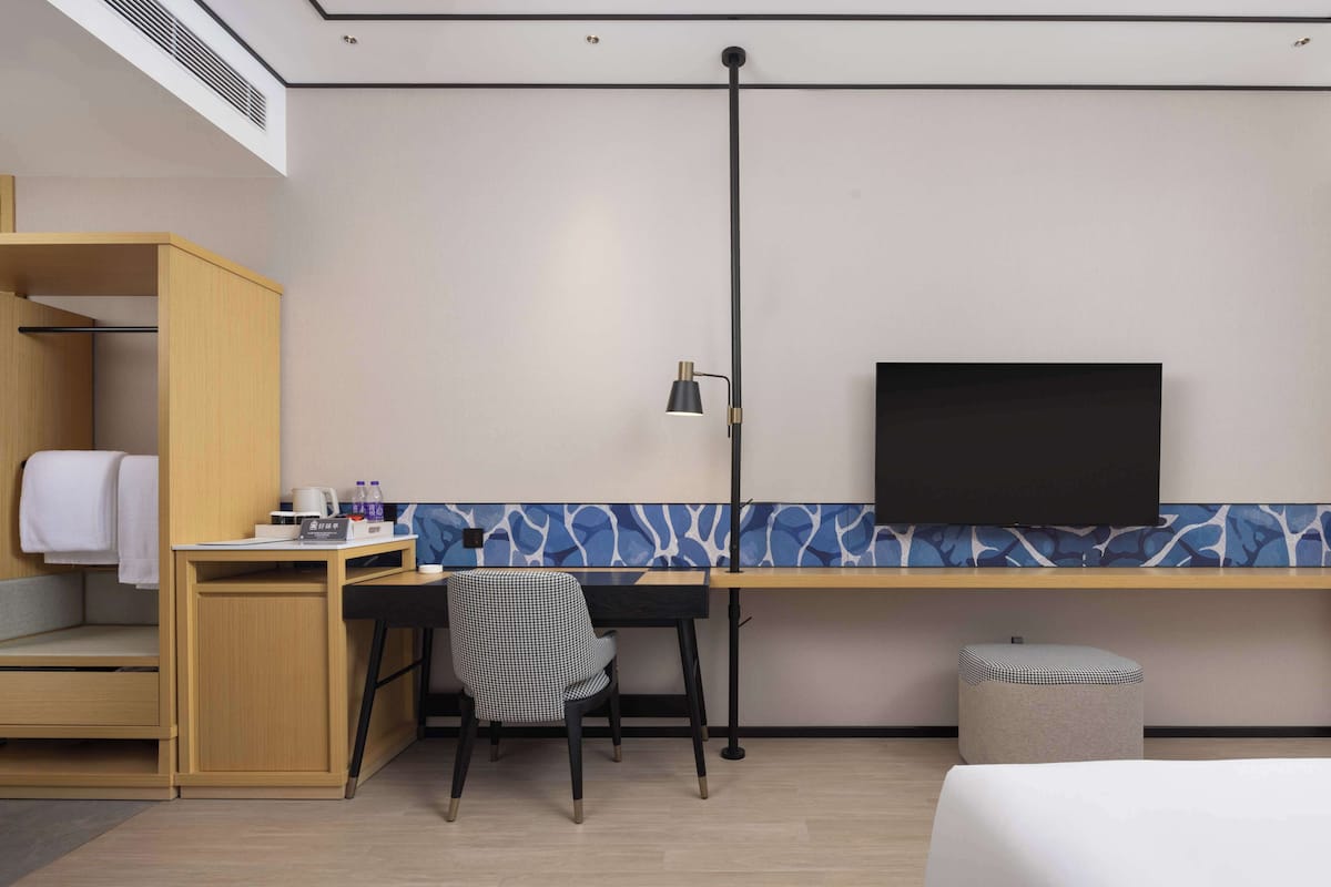 Habitación de hotel con cama, escritorio con silla, televisor, armario y un taburete pequeño.