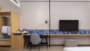 A hotel room with a bed, a desk with a chair, a television, a wardrobe, and a small stool.