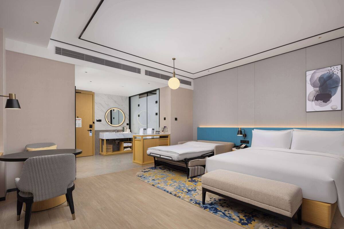 Una habitación de hotel moderna con cama, mesita de noche, silla, escritorio y baño con espejo y lavamanos.