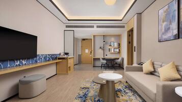 A modern hotel room with a flat-screen TV, a sofa, a dining area, and a small kitchenette.
