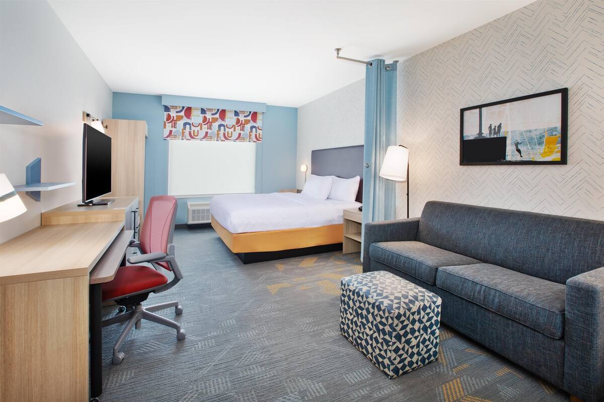 Habitació d'hotel moderna amb un llit, un escriptori amb ordinador, un sofà i un otomà estampat.