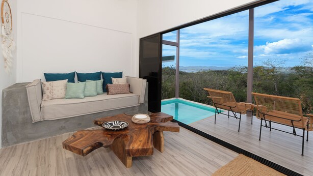 A modern living room with a sofa, coffee table, and a view of a swimming pool and natural landscape. - Essence of Honey Villas & Safari Boutique - Villa # 1 (Provincia de Guanacaste)
