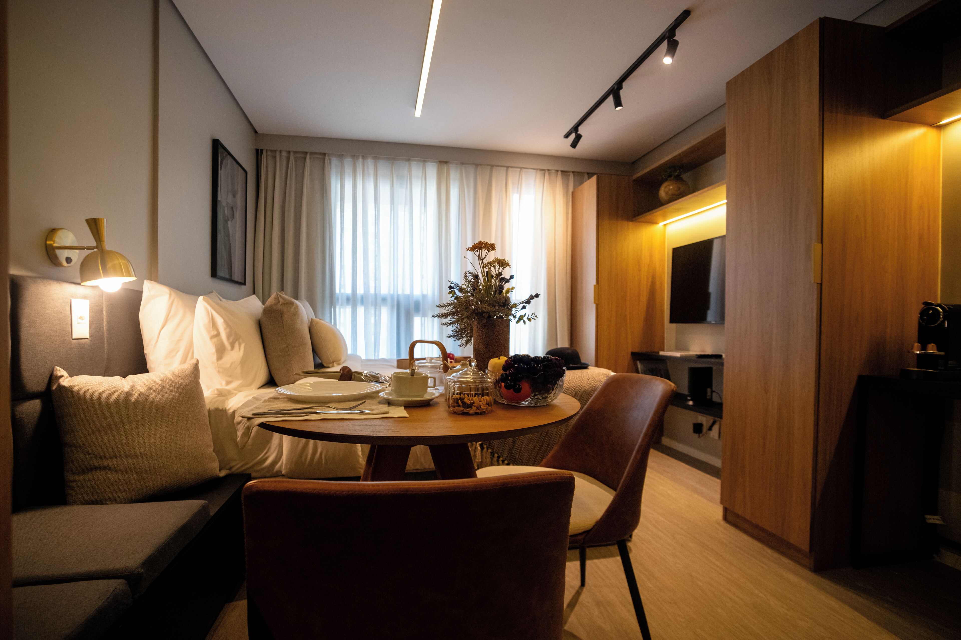 A modern hotel room with a sofa, dining table, and chairs. There is a TV, a plant, and a window with curtains.