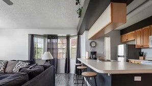 A modern living room with a dark sofa, a coffee table, and a lamp. There is a kitchen area with wooden cabinets and a countertop. - condo2/2 pretty Close to Disney (Kissimmee)