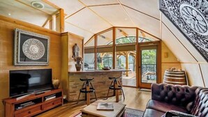 Amplia sala de estar con un televisor de pantalla plana grande, un sofá de cuero y una mesa de centro. Se observa una zona de cocina con taburetes y una encimera de piedra.
