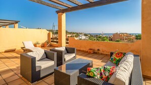 Terraço no telhado com móveis de vime, uma mesa de centro de vidro e vista para o mar e a paisagem urbana.
